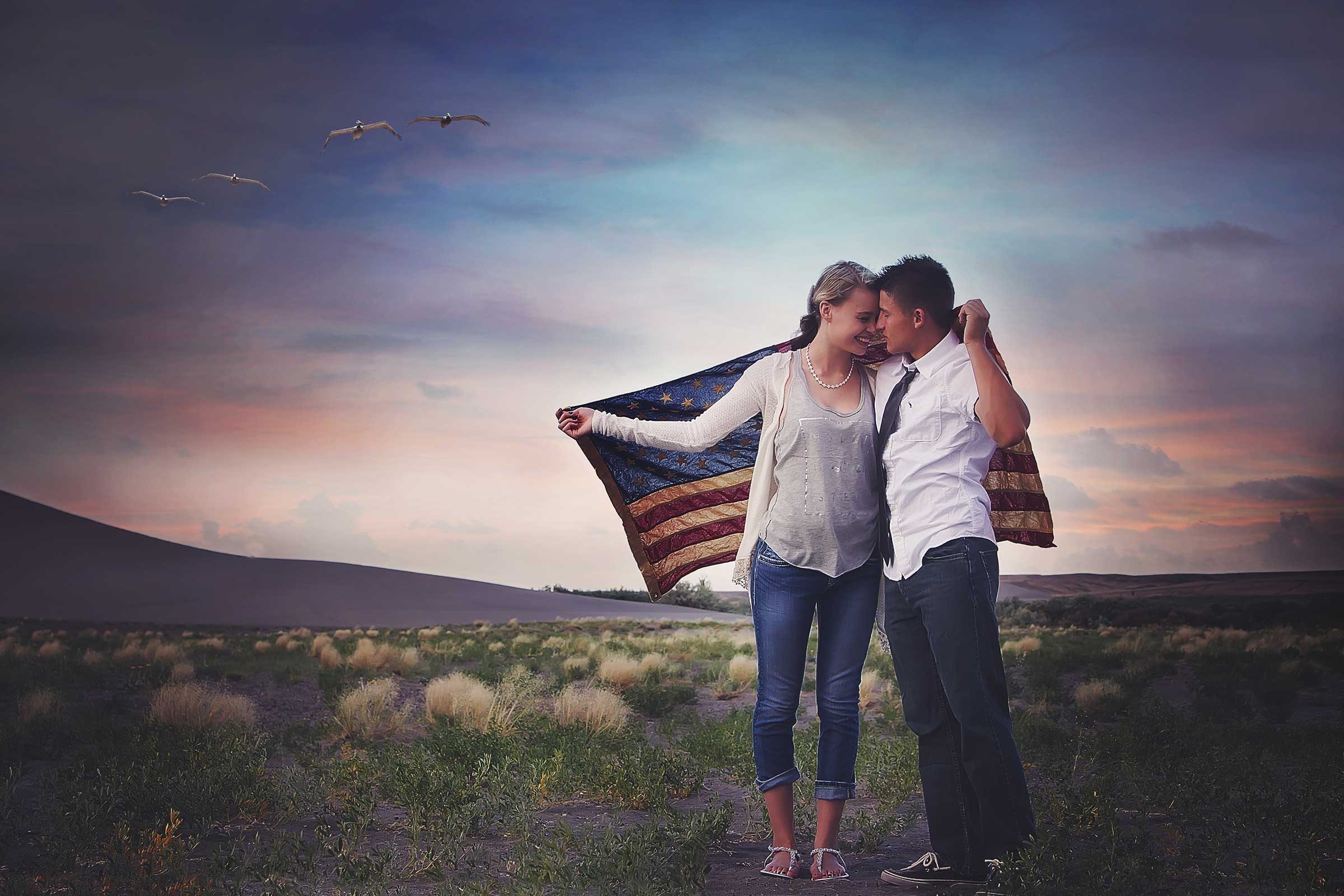 portrait of a couple holding the american flag around themselves standing in a landscape at sunset