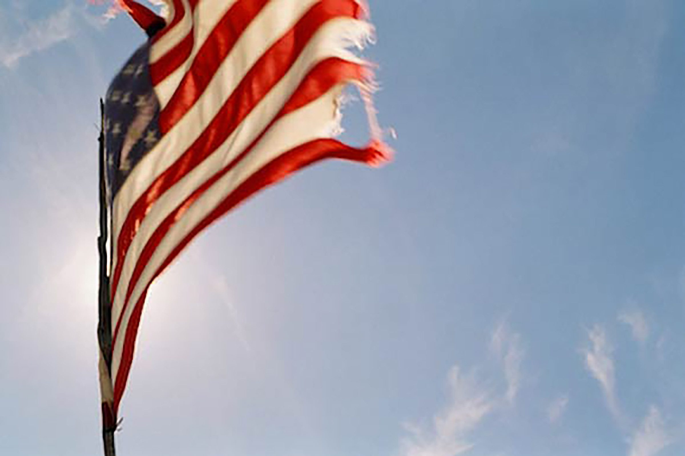 a wind tattered american flag lit from behind by the sun waves vibrantly against the blue of the sky