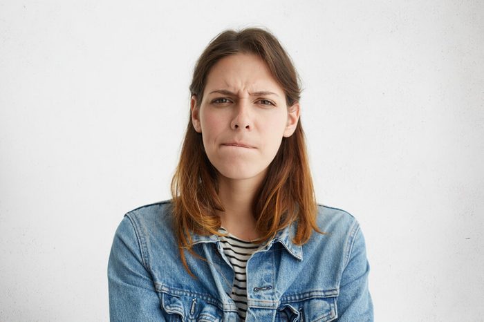 Headshot of indecisive confused young European woman in denim wear pursuing lips, her look expressing doubt and uncertainty as she has to come up with best solution while dealing with problem