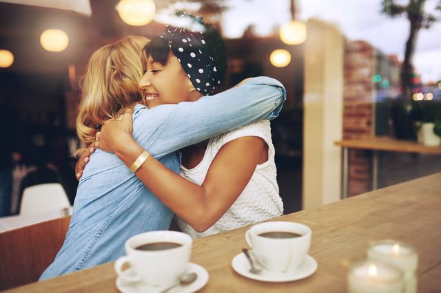 Two multi ethnic affectionate girl friends embracing as they sit in a coffee shop enjoying a cup of coffee together