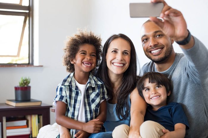 Multiethnic family taking a selfie photo while sitting on sofa at home. Laughing mother, father and sons take a selfie with smartphone. Happy parents sitting on couch with kids.