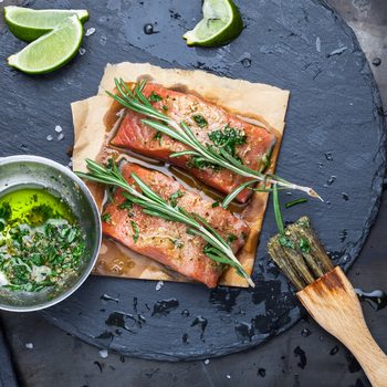 Pieces of raw salmon oiled marinade with spices, lemon, spices and olive oil with brush on slate stone on a dark metallic background. View from above. Preparation for cooking fish food. Salmon steak.