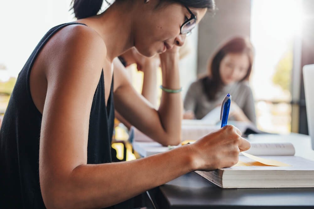 Close up shot of young female students writing notes with classmates reading in background. Students studying in university library.