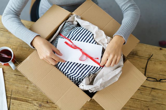 Woman packaging a Christmas gift for posting tying a red ribbon around an envelope on a giftwrapped box in a cardboard carton, high angle view