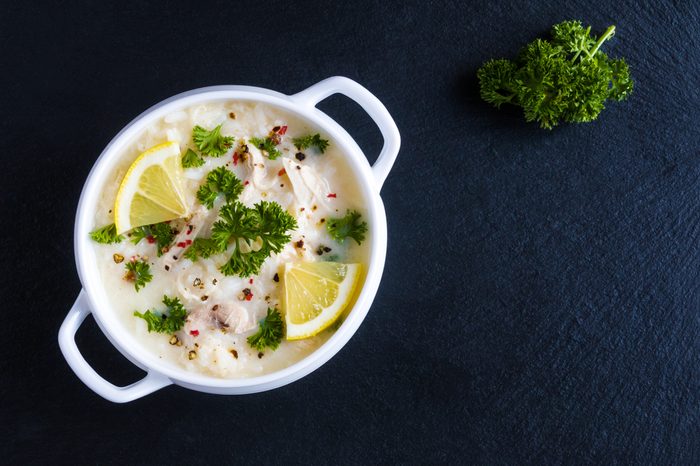 Avgolemono, chicken soup with egg-lemon sauce, rice and fresh parsley leaves in white bowl on black stone background. Top view.