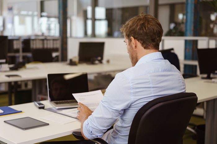 Businessman sitting at a table reading a document in a large open plan office, view from behind over his shoulder