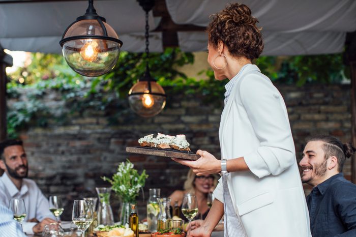 Pretty Caucasian smiling woman putting a plate with meal on dining table at backyard celebration.