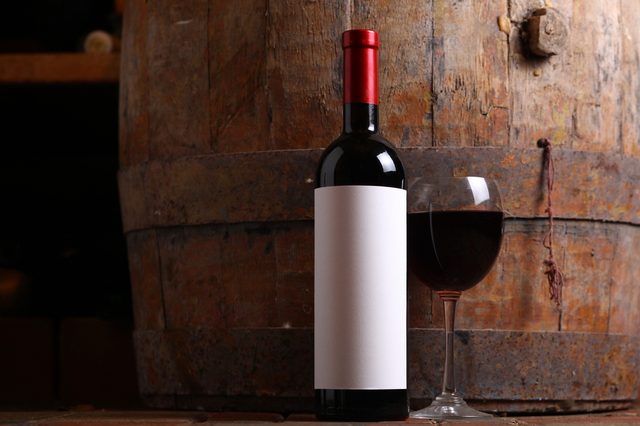 Bottle of red wine with a blank label and glass standing on cellar floor with a wooden wine barrel in the background