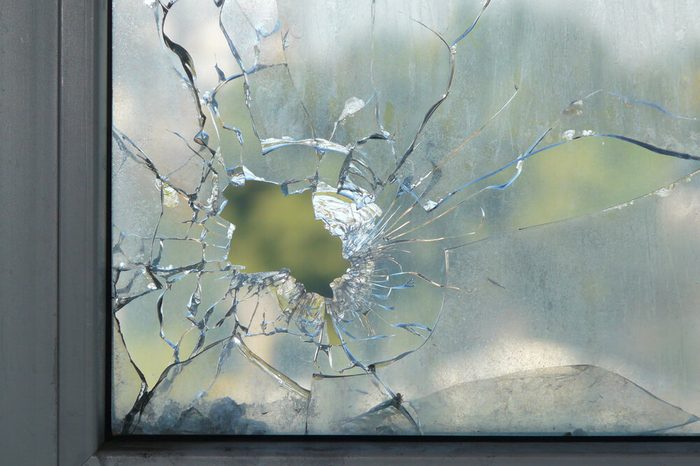 Hole in the window glass by a bullet shot during war shooting. Cracks spreading around the hole. Green leaves of trees blurred by the glass. Dirty window frame. View of a street from the inside.