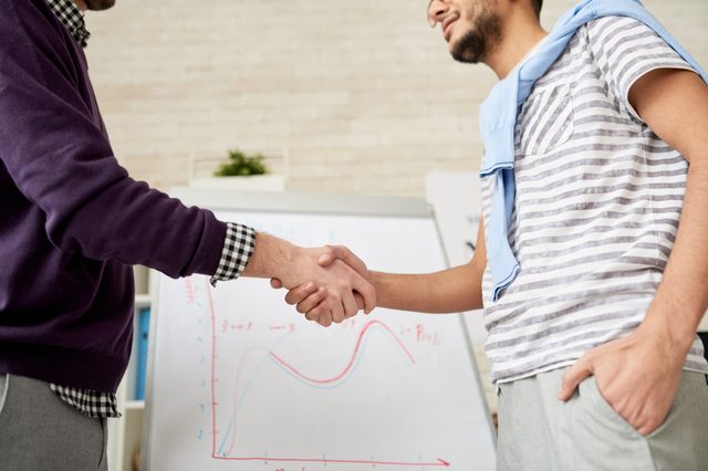 Mid-section portrait of two unrecognizable businessmen shaking hands against whiteboard with statistics graph in modern office