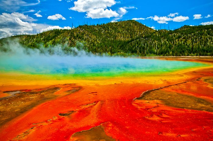 Beautiful cerulean geyser surrounded by colorful layers of bacteria, against cloudy blue sky.