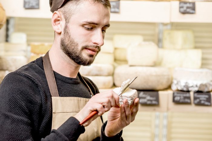 Portrait of a handsome seller in uniform cutting young cheese in front of the store showcase full of different cheeses