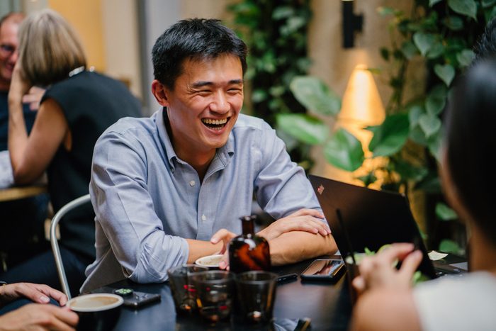 A group of young and energetic Chinese Asian coworkers sit around a table and have a light hearted business discussion. They are smiling and laughing as they talk and have a conversation.