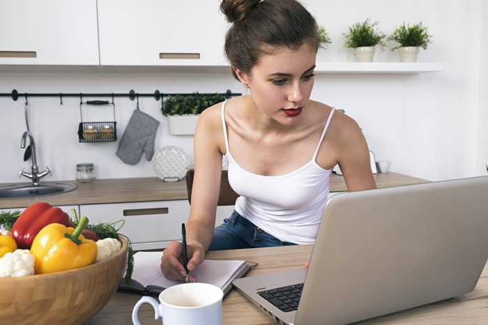 Portrait of brunette with red lips writing down information in notebook from laptop.Kitchen interior.At home