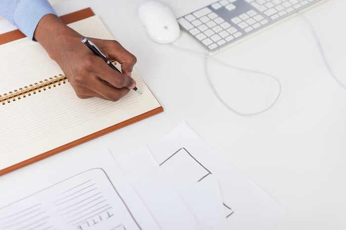 Black man hand writing in notebook. Computer mouse and keyboard near him on table. Concept of office work and decision making. Top view