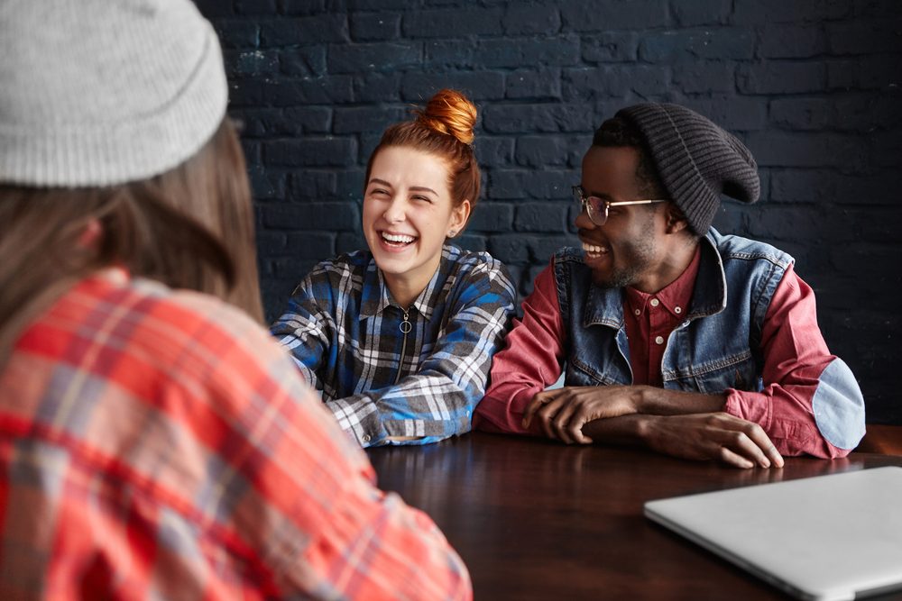 African man wearing hipster black hat and glasses and pretty girl with ginger hair talking to their common female friend in red checkered shirt, having nice time together during breakfast at cafe