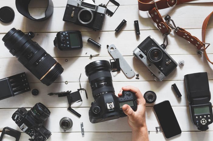 Man hand Holds Digital Camera On Table Next To Lenses And Accessories On White Wooden Background. Top View