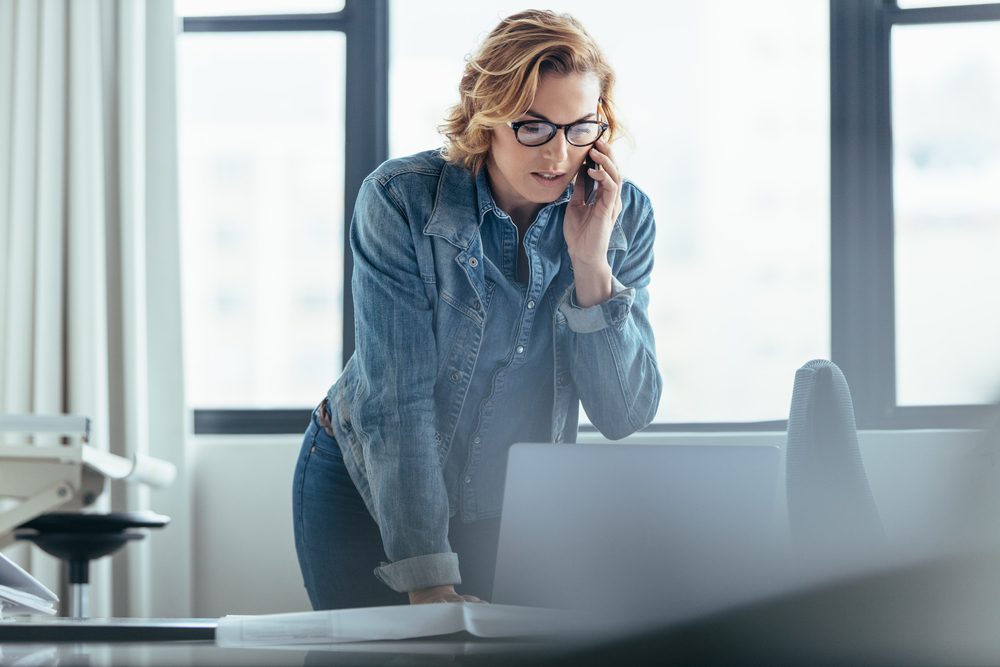 Businesswoman talking on cellphone and looking at laptop. Female executive working in office.