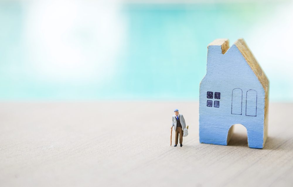 Miniature old man carry grocery bag with blue wooden miniature house over blurred blue background, outdoor day light