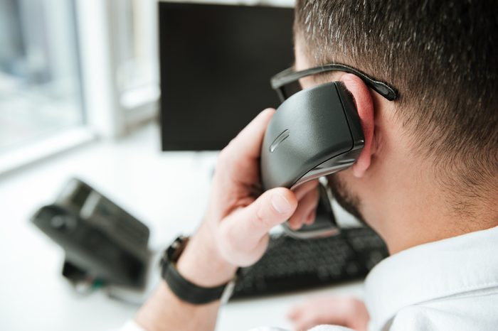 Back view of man in white shirt and eyeglasses which sitting by the table with computer and talking on phone. Close up view