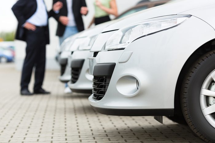 Two men man and one woman stand behind a row of silver-metallic cars on a parking place on yard of dealer