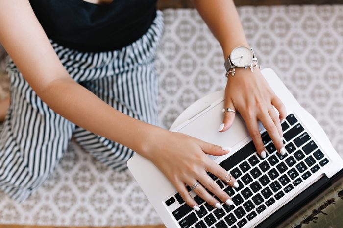 Woman with tanned skin typing on keyboard while standing on her knees. Girl in trendy silver accessories using laptop sitting on carpet with ornament.