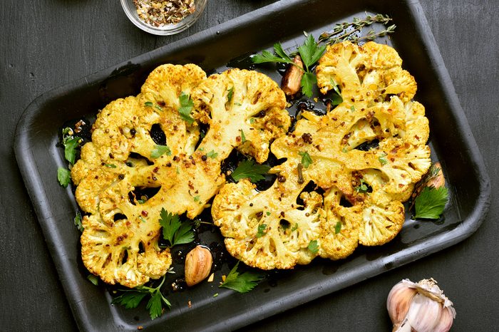 Baked cauliflower steaks with herbs and spices on baking sheet over black stone background.