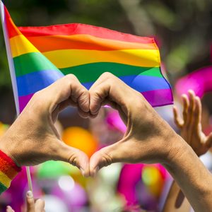 Supporting hands make heart sign and wave in front of a rainbow flag flying on the sidelines of a summer gay pride parade