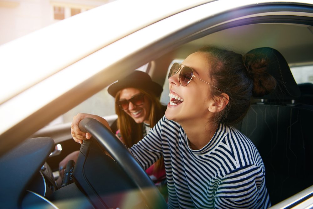 Laughing young woman wearing sunglasses driving a car with her girl friend , close up profile view through the open window