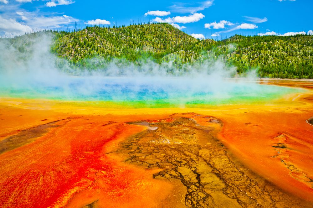 Beautiful cerulean geyser surrounded by colorful layers of bacteria, against cloudy blue sky.