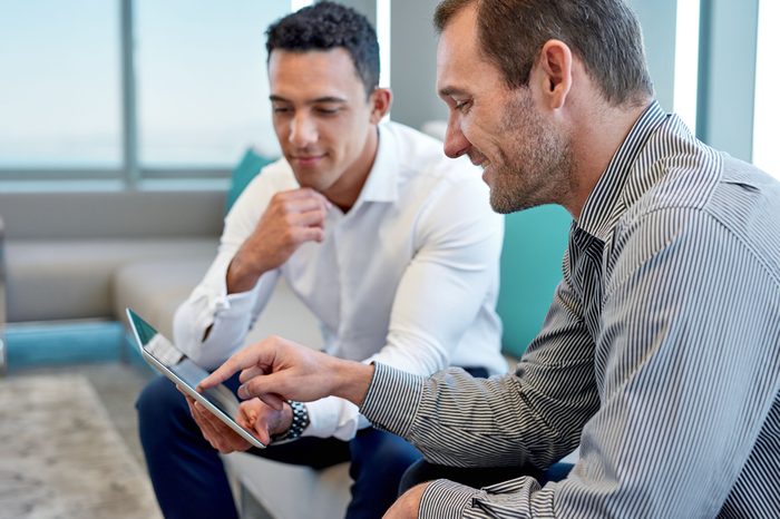 Two smiling male work coworkers sitting on a sofa in a modern office talking together over a digital tablet