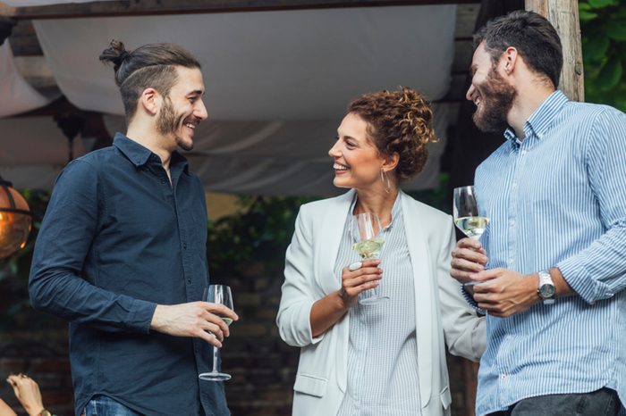 Pretty elegant Caucasian woman chatting with two men and drinking wine at outdoor celebration.