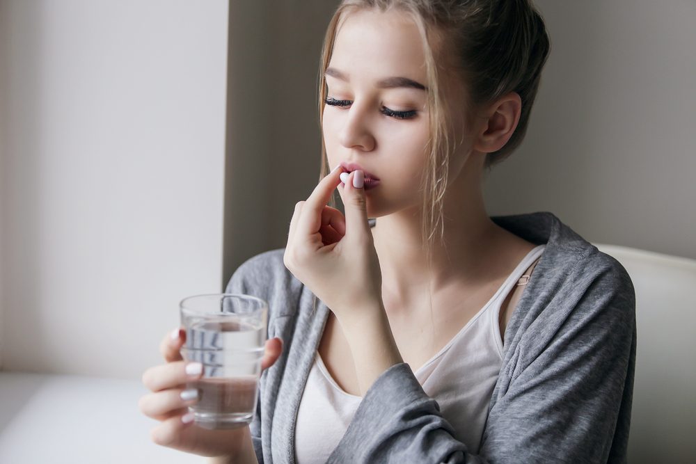 Young beautiful girl or woman to take a pill with a glass of water near the window in white shirt and grey robe