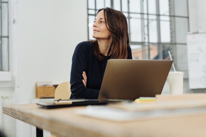 Businesswoman sitting watching to the side with a smile while leaning on her desk with a laptop computer in a low angle view