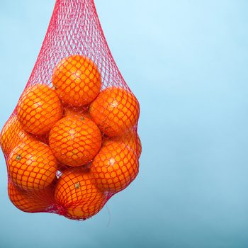 Mesh bag of fresh oranges healthy tropical fruits from supermarket on blue. Food retail.