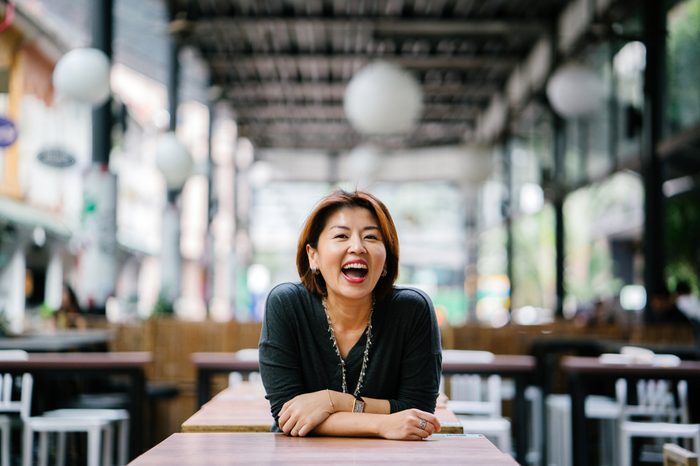 Portrait of an elegant mature Asian woman laughing during the day. She's dressed professionally and leaning against a table in a coworking space, cafe or office in Asia. 