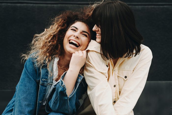 Portrait of two attractive girls dressed in jeans blue and yellow jackets having fun and laughing against a black wall.