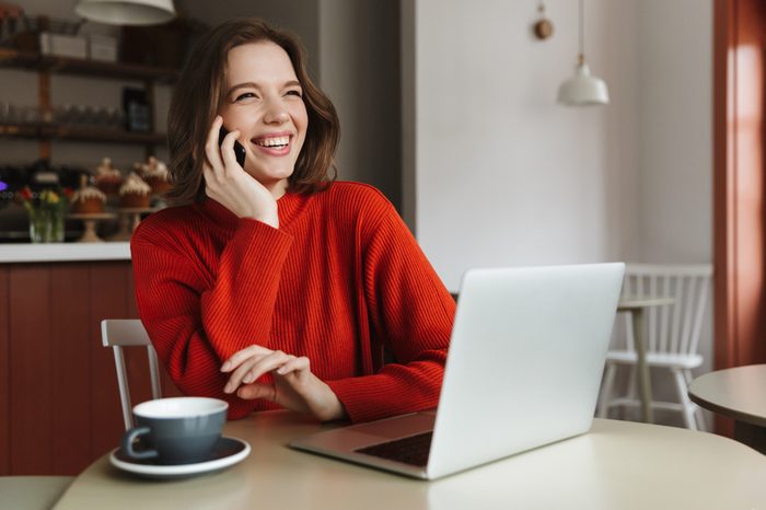 Image of happy caucasian woman 20s laughing and talking on smartphone while using laptop in cafe