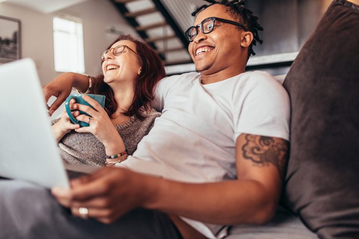 Cheerful multi-ethnic couple relaxing together on sofa with laptop and cup coffee. Smiling man and woman sitting on couch at home.