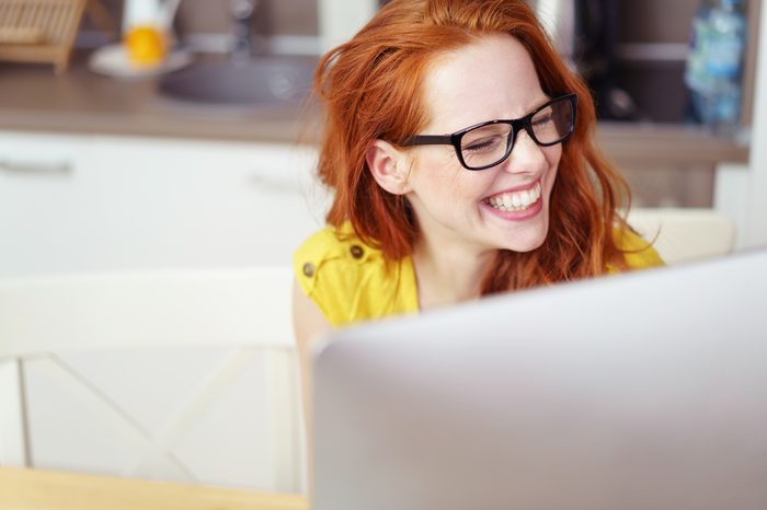 Head and Shoulders of Young Woman with Red Hair Wearing Eyeglasses and Laughing Joyfully While Working on Computer with Over-Sized Monitor at Home in Kitchen