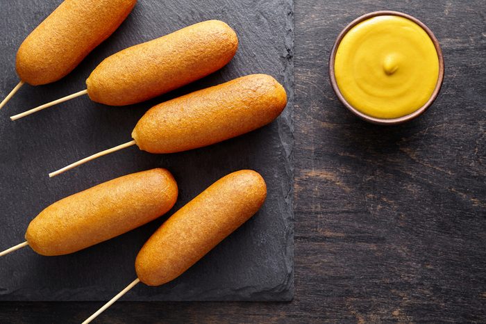 Corn dog traditional American corndog street junk food deep fried hot meat sausage snack with yellow mustard treat coated in a layer of cornmeal batter on stick on black rustic desk.