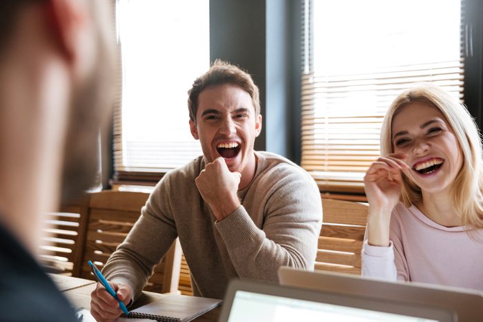 Photo of three young laughing colleagues sitting near coffee while work with laptops and notebook. Coworking.