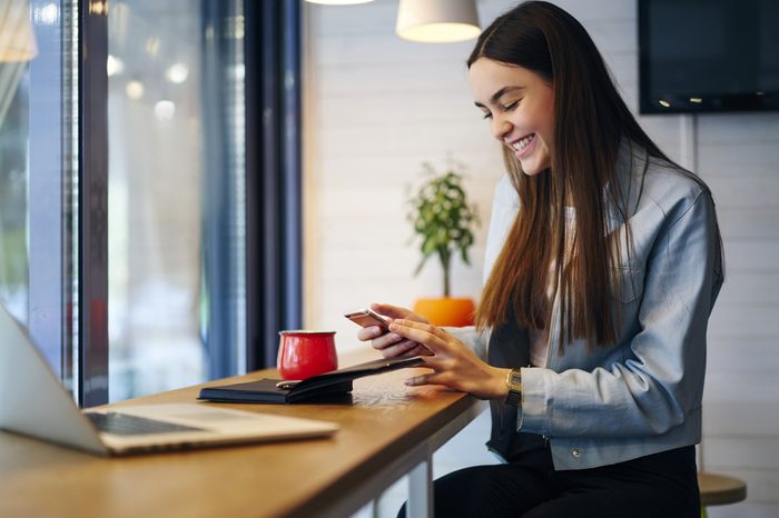 Positive young woman reading funny sms message on modern smartphone and laughing while sitting in cafe interior at table with laptop computer and notepad.Smiling female chatting in social networks