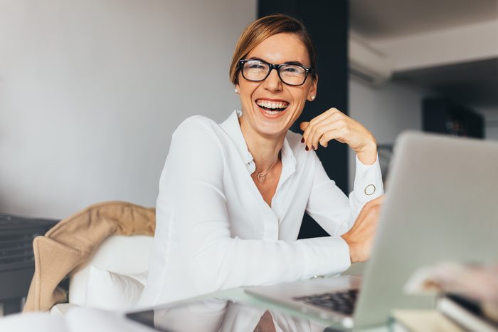 Business woman in spectacles sitting at her desk in office and laughing. Woman sitting in office with laptop computer on her desk.