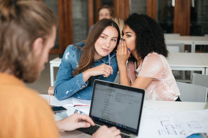 Portrait of two smiling beautiful girls sitting in classroom and gossiping while young man working on his laptop. Pretty girl with dark curly hair saying secret to her friend in office