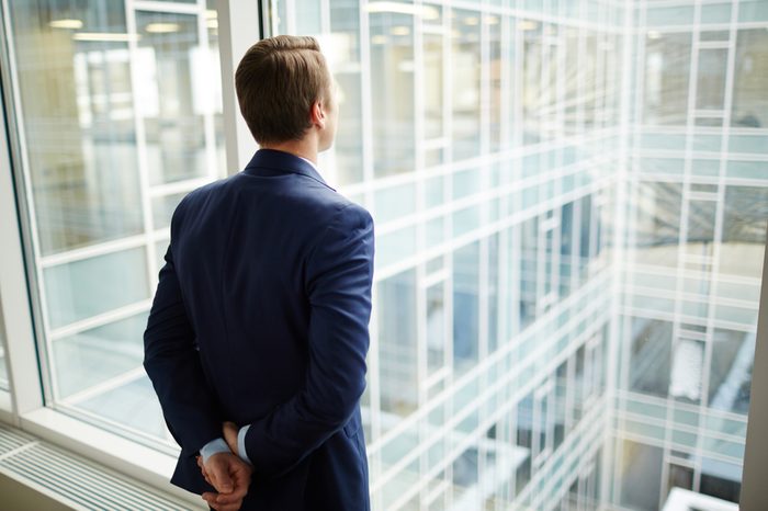 Rear view of elegant businessman standing by window of new office and looking through it