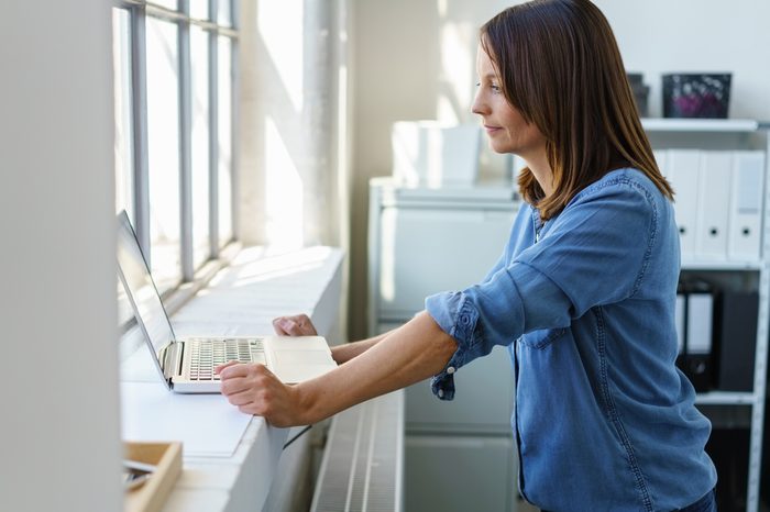 Businesswoman standing at a large window sill using her laptop computer in the bright daylight in a profile view