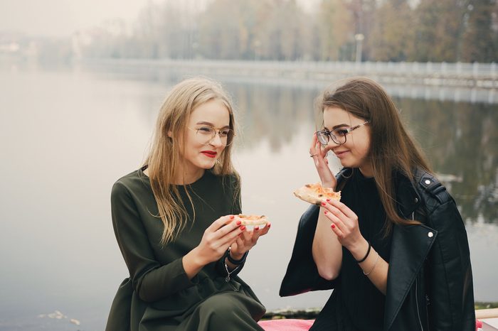 two young and stylish girls walking in the autumn park near water and eating pizza