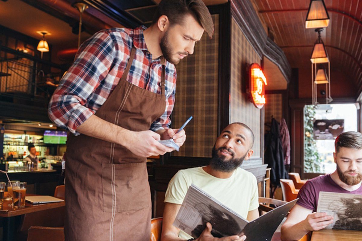 Serious concentrated young waiter standing and taking an order from two bearded handsome men in cafe