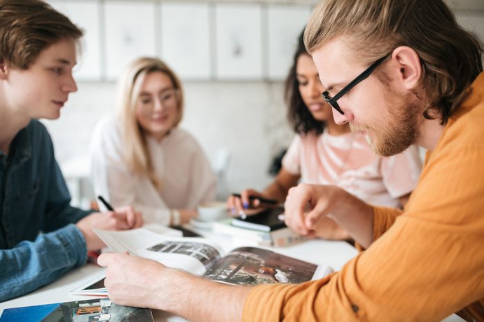 Portrait of young man with blond hair and beard thoughtfully looking in journal with friends on background. Cool boy in glasses sitting in office and working with friends 